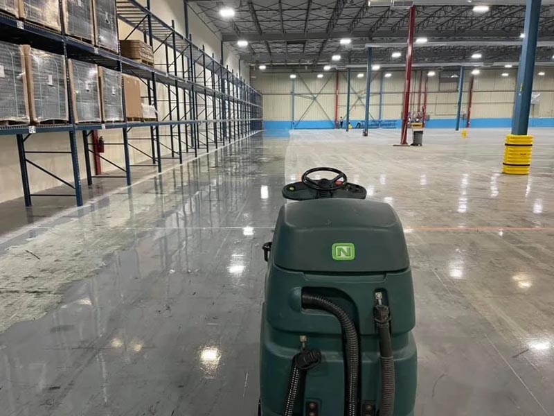 A large warehouse with high shelves partially filled with boxes. A floor-cleaning machine is in the foreground on the shiny, polished concrete floor. The space is well-lit with ceiling lights and has a blue and white color scheme.