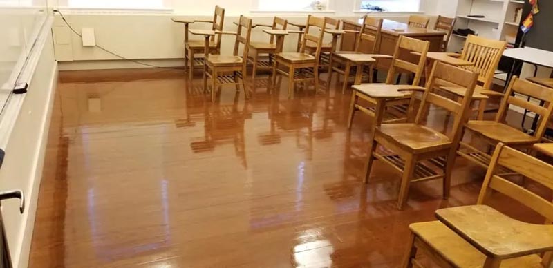 An empty classroom with polished wooden floors, featuring wooden chairs with attached desks arranged in rows. Sunlight streams through windows in the background.