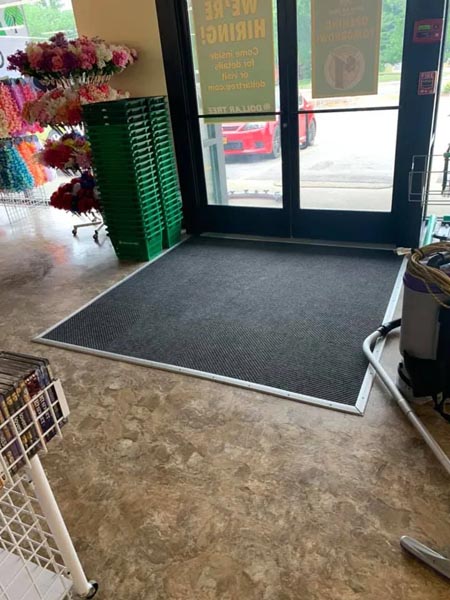 Store entrance with glass double doors and a dark grey floor mat. Stacks of green shopping baskets are on the left and displays of colorful artificial flowers are nearby. A red car is visible outside.