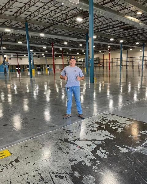 A man wearing a gray T-shirt, blue jeans, and a cap stands in a large, empty warehouse with a high ceiling and multiple support pillars. The floor is shiny and reflective.