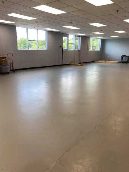 A large, empty room with smooth, gray flooring and overhead fluorescent lighting. The walls are light-colored with several windows letting in natural light. A red hand truck and wooden pallet are near one wall.