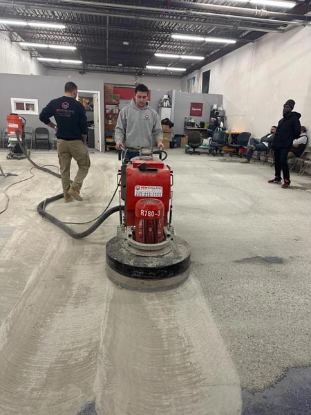 A person operates a red floor grinding machine on a concrete floor in an industrial space. Two other people stand nearby, with one seated in the background. The area has unfinished walls and a glimpse of an office in the corner.