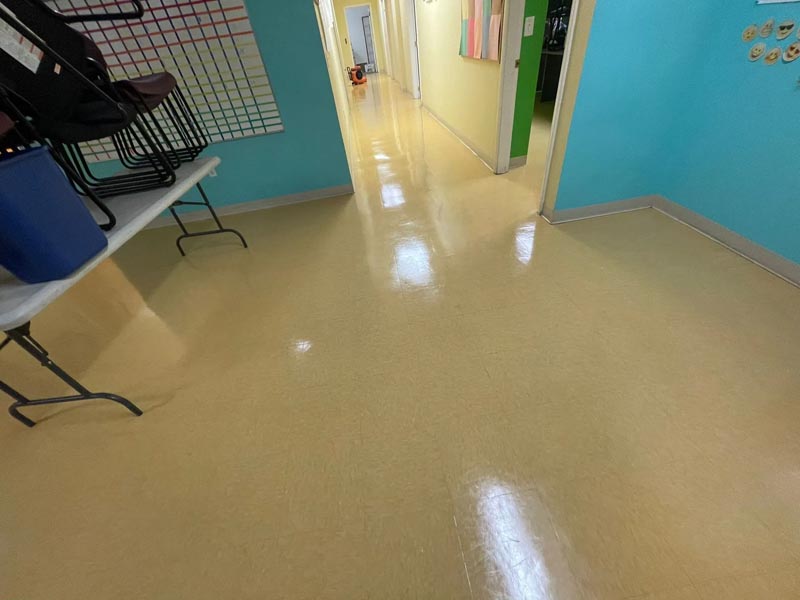 A brightly lit hallway with yellow floors and colorful walls. Chairs are stacked on a folding table to the left, and a chart is visible on the wall. The corridor leads to various rooms and is empty of people.