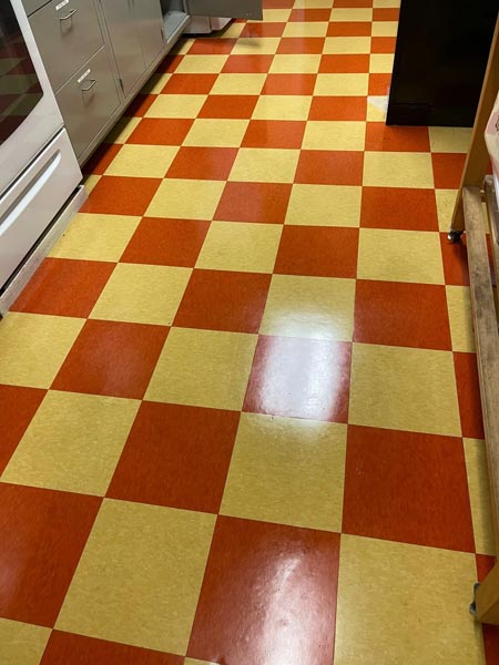 A kitchen floor with a shiny checkered pattern in red and yellow tiles. The floor is surrounded by white and silver kitchen cabinets and appliances.