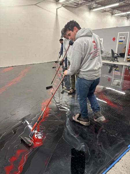 Workers in casual clothing spread a glossy, dark-colored epoxy over a large floor using long-handled rollers. The scene takes place in a spacious indoor area with high ceilings and partially painted walls.