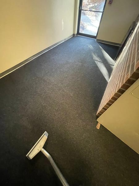A vacuum cleaner is being used on a dark carpeted floor in a room with light-colored walls. Sunlight streams in through a window, casting shadows across the floor. A mattress leans against the wall on the right.