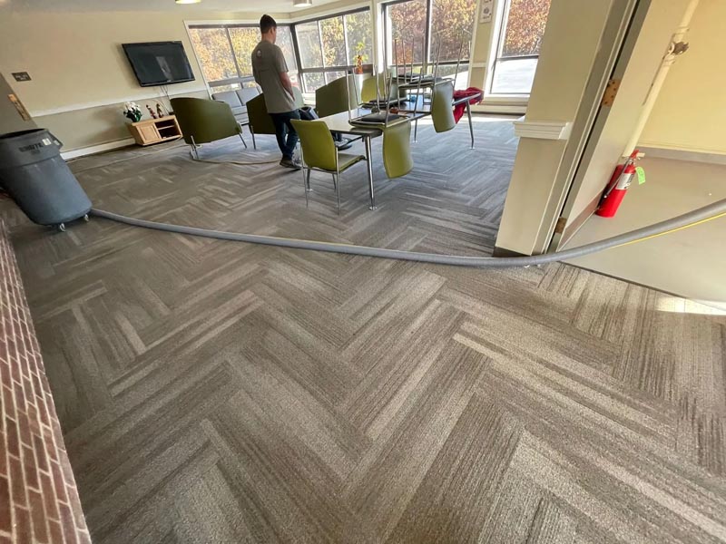 A person walks across a room with a patterned carpet. Chairs and a table are arranged near large windows. A vacuum hose is stretched across the floor, and a fire extinguisher is mounted on the wall.