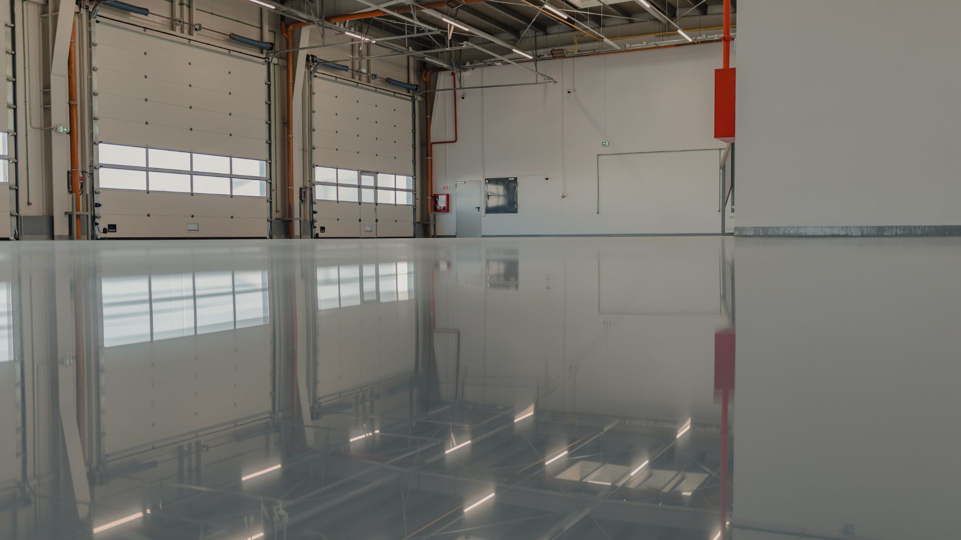 An empty warehouse with polished concrete floors reflecting the ceiling. Large bay doors line one wall, and there are pipes and a fire extinguisher box on the walls. The space is lit with fluorescent lights.