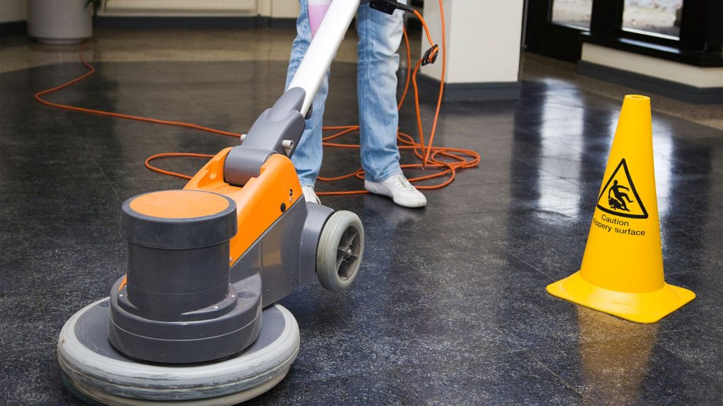 A person cleans a shiny black floor using an orange and gray floor polisher. Next to them, a yellow caution sign warns about a slippery surface. An orange extension cord is visible on the floor.