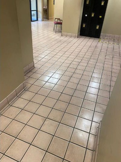 A hallway with shiny, light-colored tiled flooring. The walls are painted beige, and at the far end, there is a stack of chairs against the wall. The hallway leads to an open door on the left.