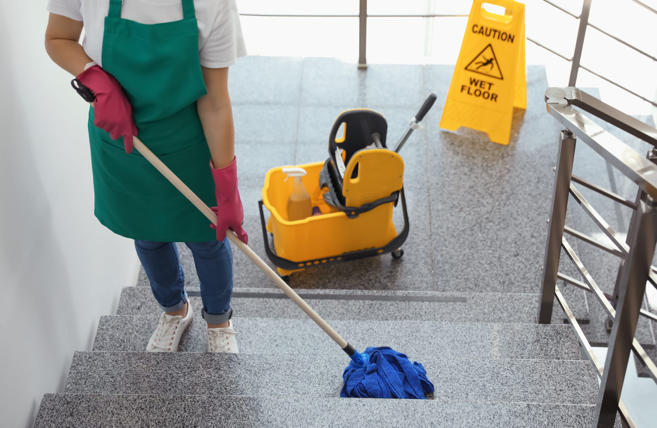 A person in a green apron and pink gloves mops a granite staircase. A yellow caution sign and a cleaning bucket with a mop wringer are at the landing. Metal railing visible on the right.