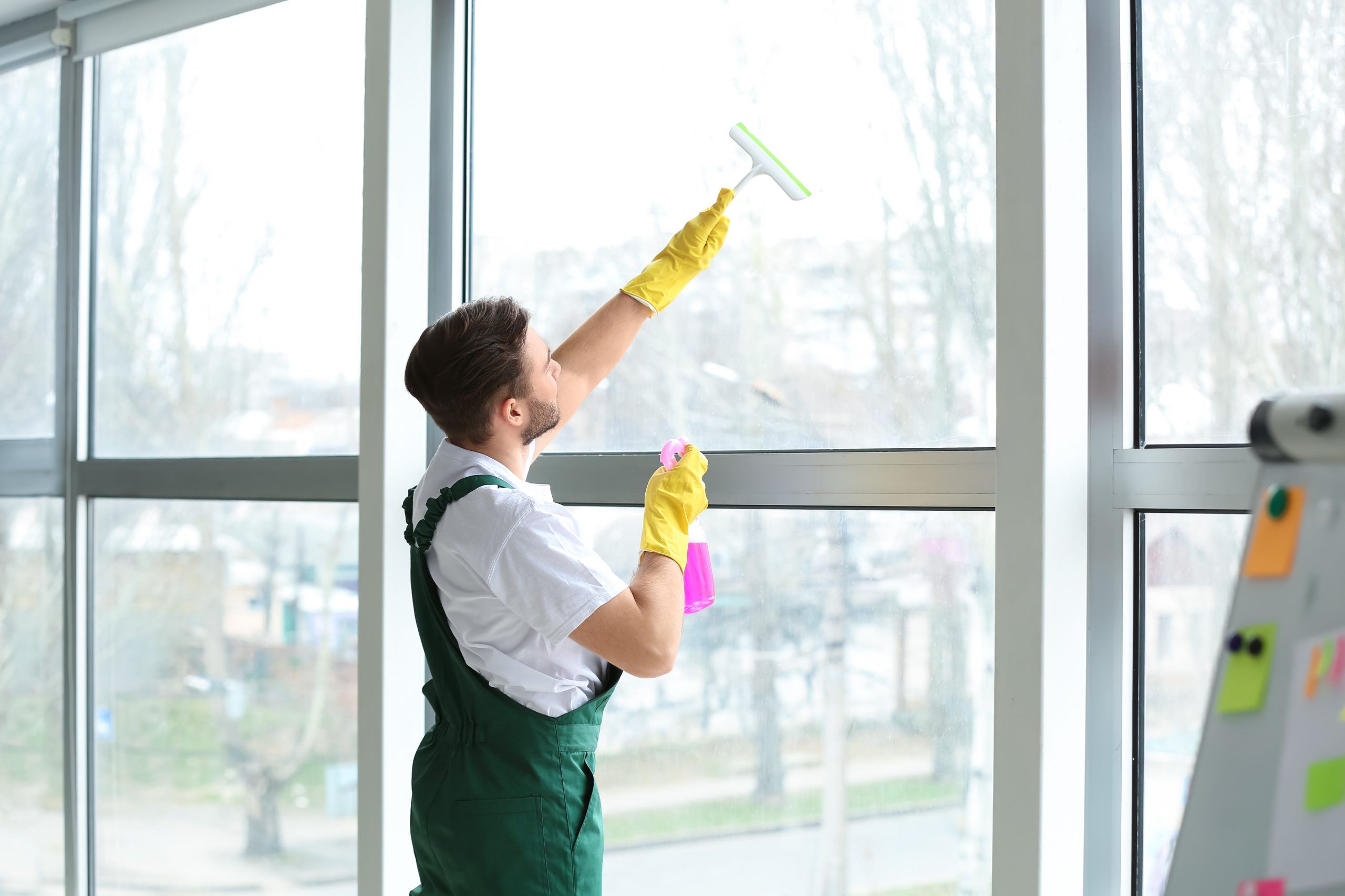 A person wearing yellow gloves and green overalls is cleaning a large window with a squeegee and a spray bottle. They are standing indoors with a blurred view of trees and buildings outside.