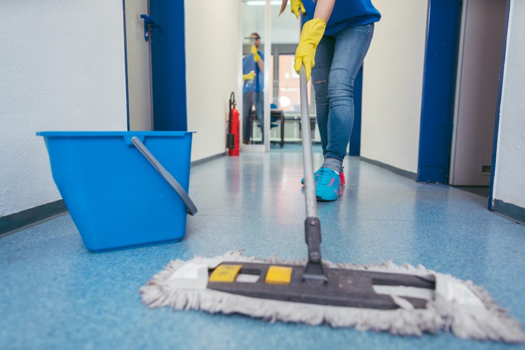 A person wearing jeans and sneakers is mopping a blue tiled hallway. They are holding a mop near a blue bucket. A fire extinguisher is visible in the background near a door.