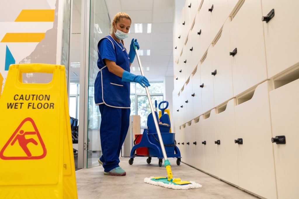 A person wearing a mask and blue cleaning uniform mops the floor in an office hallway. A yellow "Caution Wet Floor" sign is visible on the left, and cleaning supplies are on a trolley nearby. White lockers line the right side of the corridor.