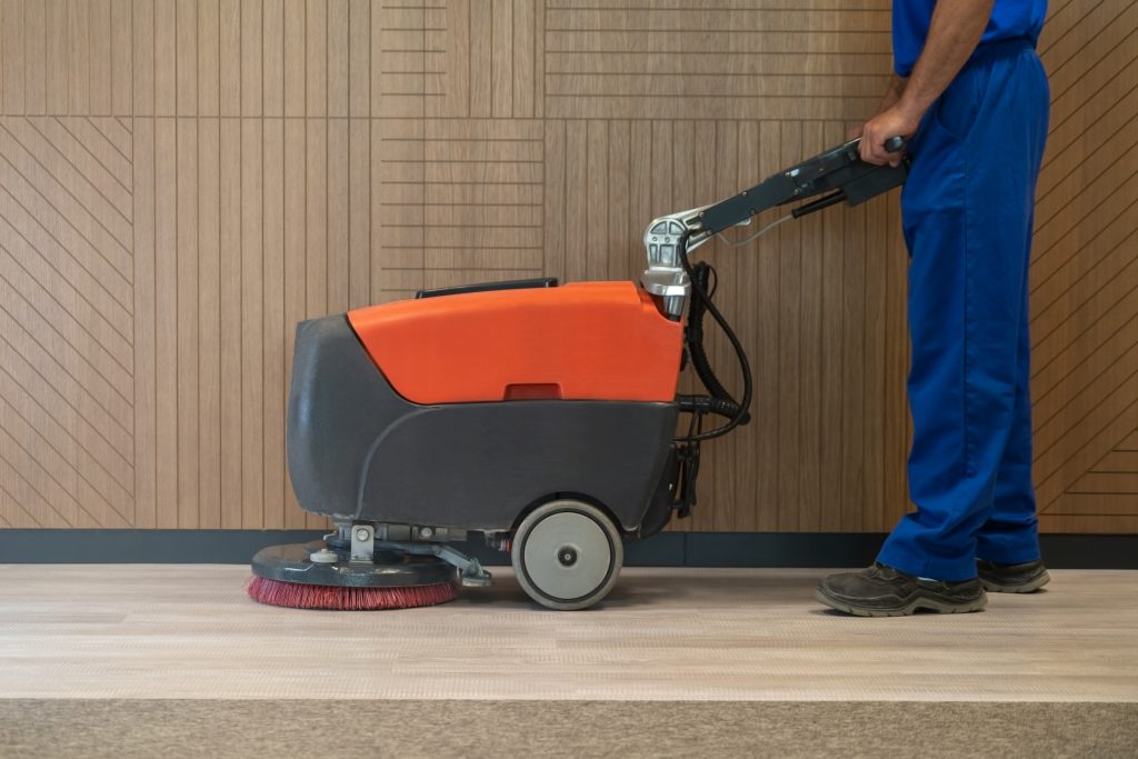 A person in blue work attire operates a floor cleaning machine with a red and black body on a wooden floor. The background features wooden paneling with geometric designs.
