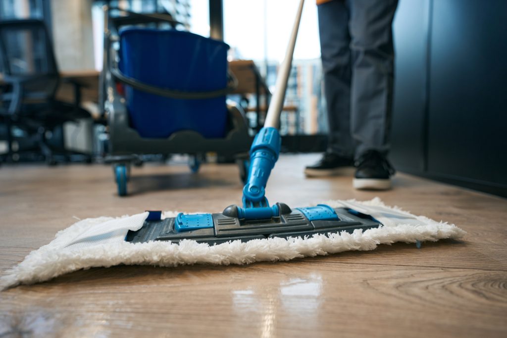 A person mopping a wooden floor in an office with a blue mop and bucket. The scene is indoors, featuring chairs and large windows in the background.