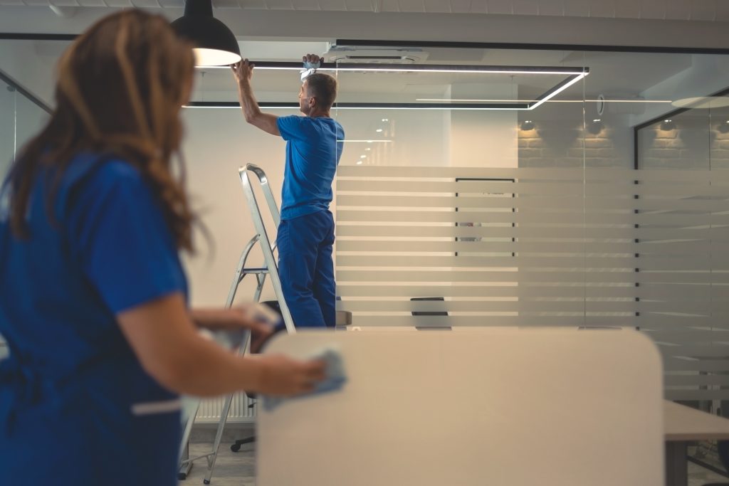 A man in a blue uniform stands on a ladder installing a light fixture in an office, while a woman in a blue uniform cleans a desk. The office has modern glass walls with striped patterns.