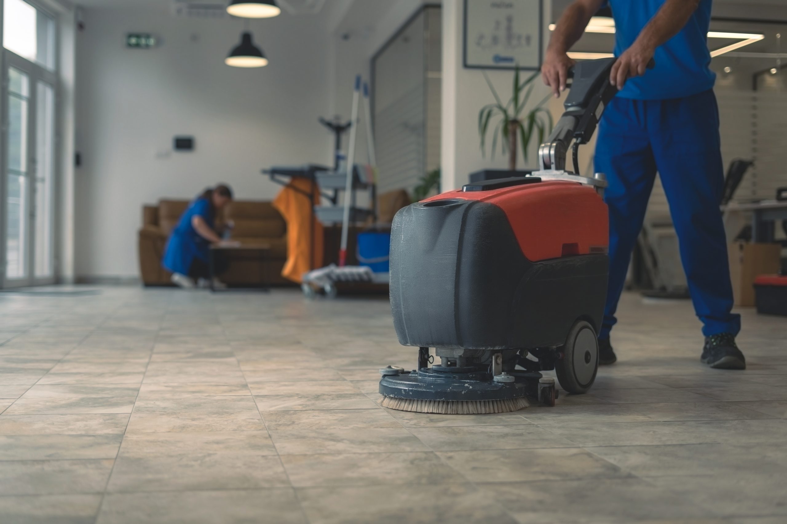 A person in blue uniform uses a floor cleaning machine on a tiled floor in a well-lit room. Another person is in the background, kneeling and cleaning a brown sofa. Equipment and cleaning supplies are visible.