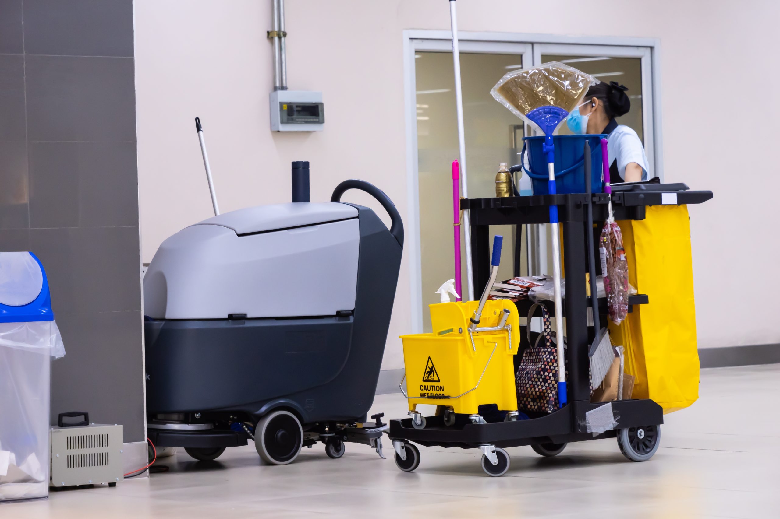 A cleaning cart with various supplies and a floor cleaning machine is in a hallway. A person is pushing the cart, and there are caution signs and mop buckets attached. The environment appears to be a clean, indoor space.
