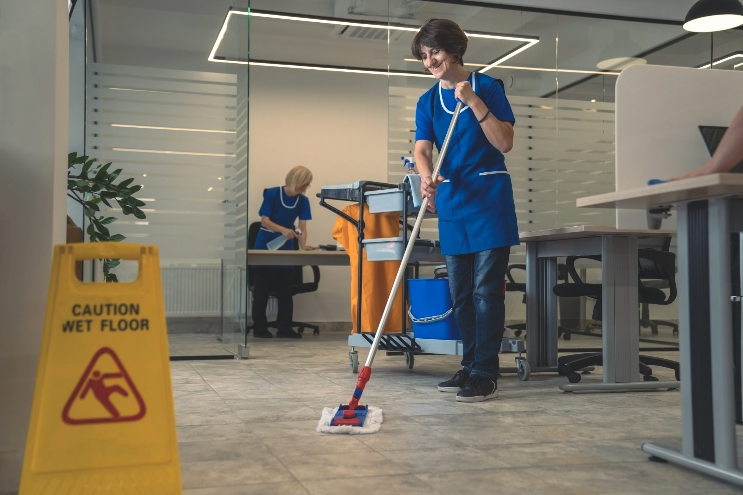 A person in a blue uniform mopping the floor in an office space. Another person is seen cleaning in the background with a cart. A yellow "Caution Wet Floor" sign is visible on the left. Desks and glass partitions are in the surroundings.