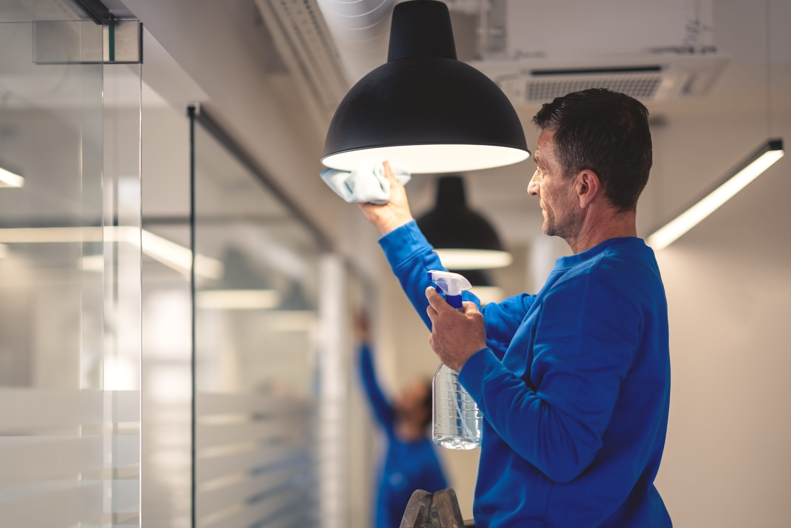 A man in a blue shirt is standing on a ladder, cleaning a black hanging lamp with a cloth and holding a spray bottle. The setting appears to be an office with glass partitions. Another person in the background cleans a glass wall.