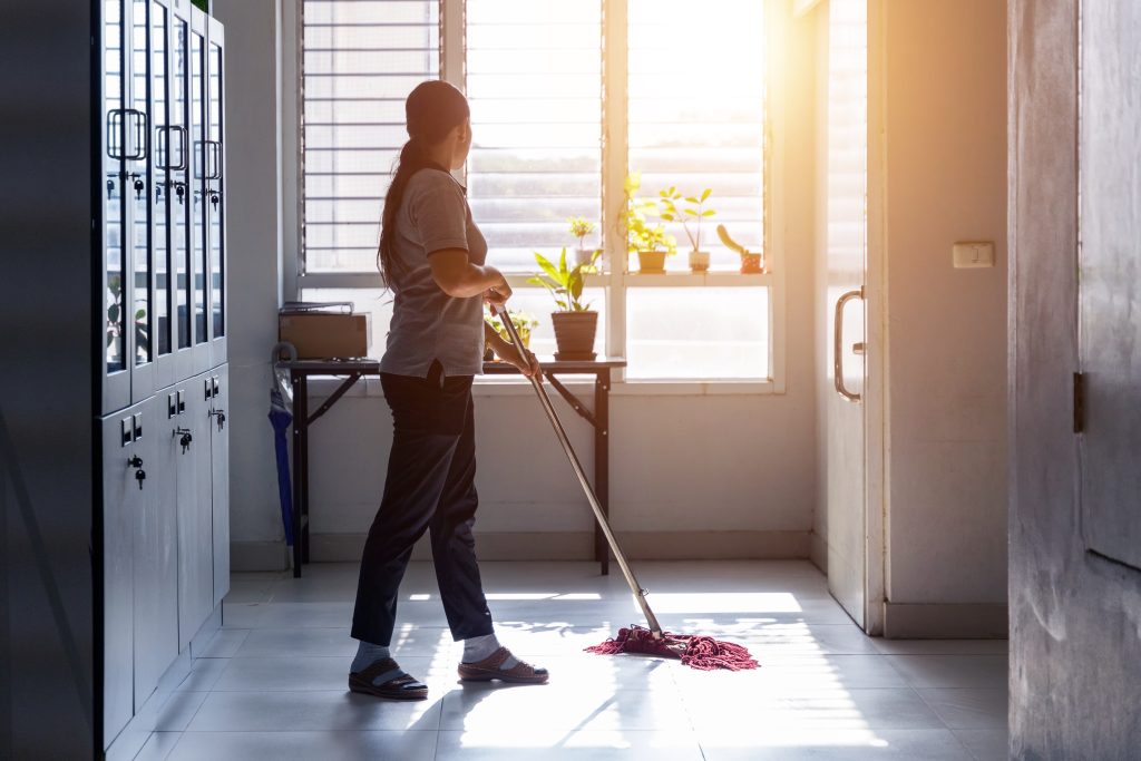 A person mops a sunlit room with potted plants on a table by the window. The room has tiled floors and large windows with blinds. Shelving units are visible on the left side. The sunlight streams in, creating a warm ambiance.
