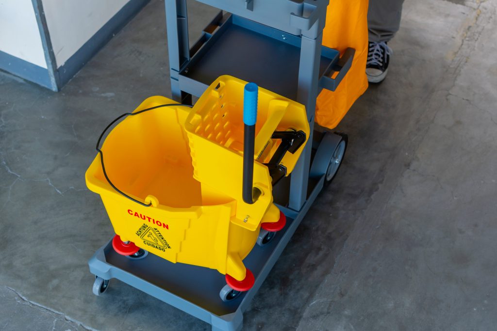 A cleaning cart with a yellow mop bucket and wringer, marked with a caution symbol, is parked on a concrete floor. An orange bucket is attached to the cart, and a person wearing sneakers is partially visible.