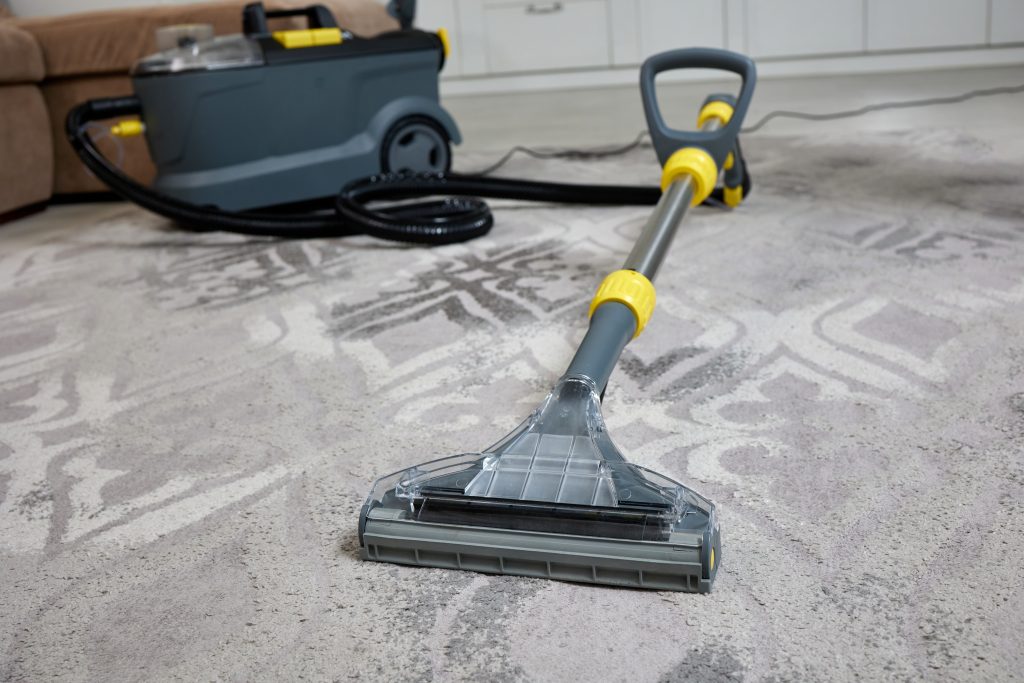 A carpet cleaner machine with a transparent nozzle is positioned on a patterned rug in a living room. A yellow handle and black hose are visible, with a couch and cabinet in the background.