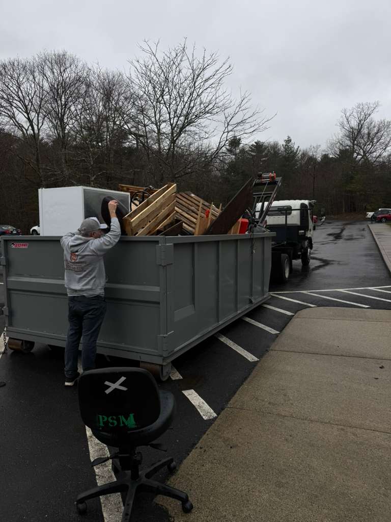 A person stands next to a large gray dumpster filled with discarded furniture and wood, while a truck is parked in the background. The scene is outdoors on a cloudy day, with bare trees and an office chair in the foreground.