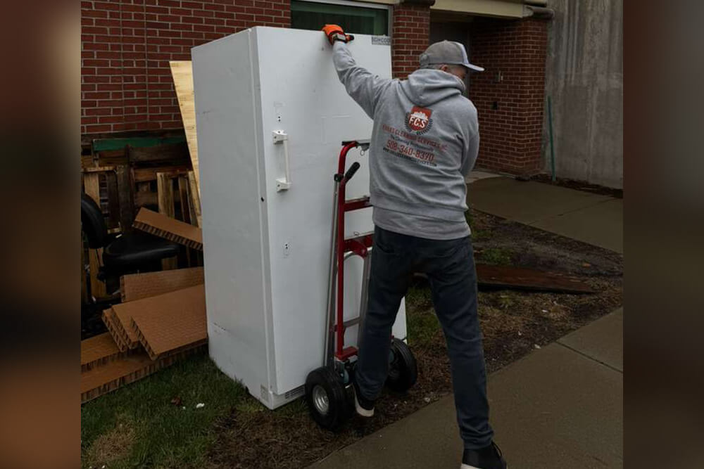 A person in a gray hoodie and cap uses a red hand truck to move a large white refrigerator outside near a brick building, with cardboard and debris in the background.