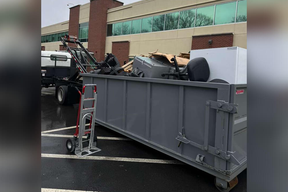 A large gray dumpster filled with discarded office furniture and equipment sits in a parking lot near a brick and glass building; a dolly and a truck are nearby on a rainy day.