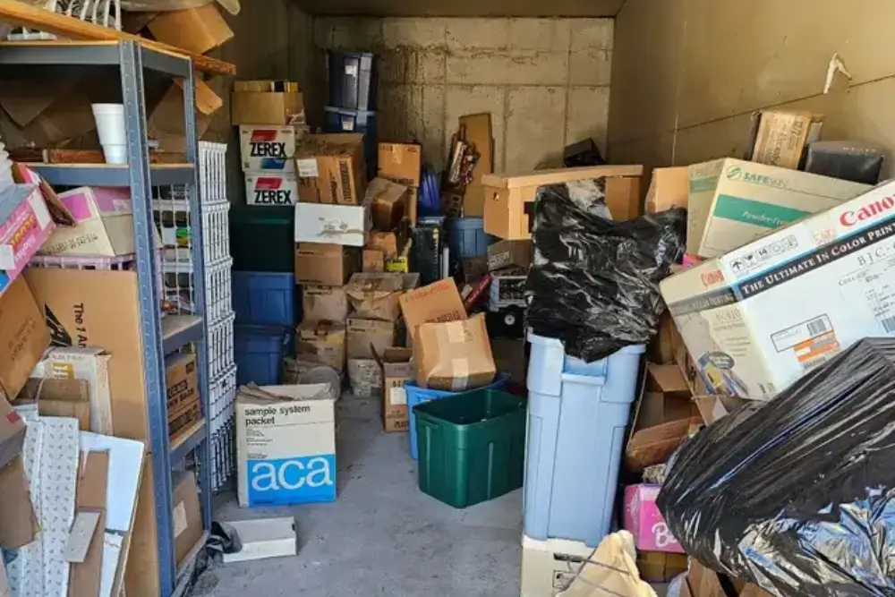 A cluttered storage unit filled with stacked cardboard boxes, bins, and various household items. Items are haphazardly arranged, making the space appear crowded and disorganized.